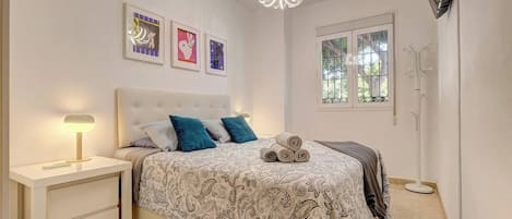 A bedroom with a bed, bedside table, lamp, artwork, and a window.