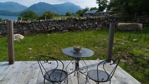 Een houten terras met twee metalen stoelen en een tafel, met uitzicht op een gazon met een stenen muur en een huis op de achtergrond.