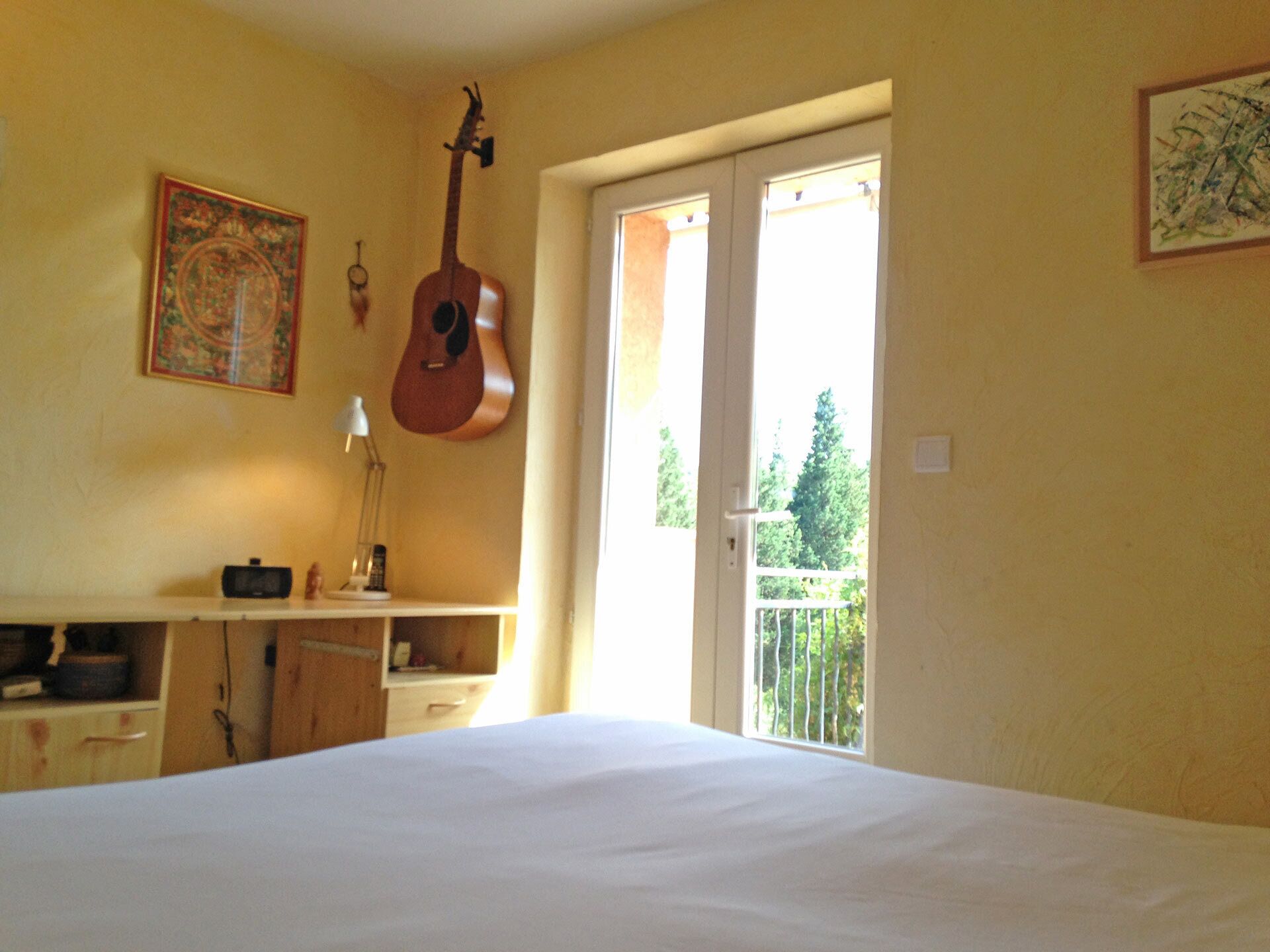 Une chambre avec un lit, un bureau avec une lampe, une guitare et un balcon donnant sur des arbres.