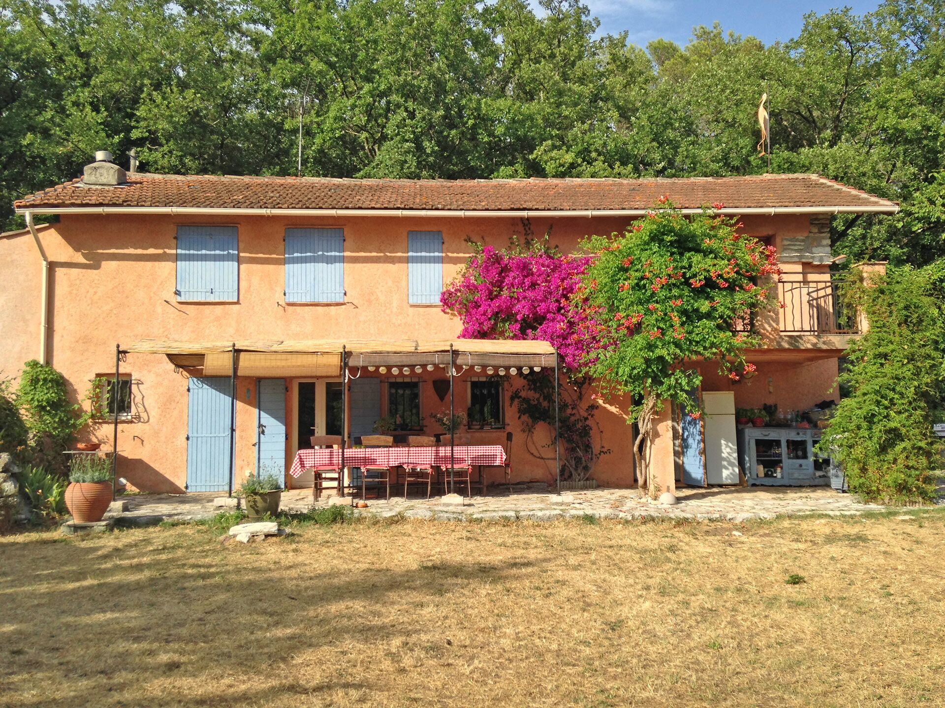Un bâtiment à deux étages doté d’un balcon, d’un espace extérieur couvert aménagé en coin salon et d’un jardin avec une grande plante en pot.