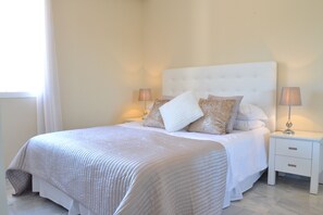 Un lit bien fait, avec une tête de lit blanche et plusieurs oreillers, flanqué de deux tables de chevet surmontées de lampes, dans une pièce au design simple et épuré.