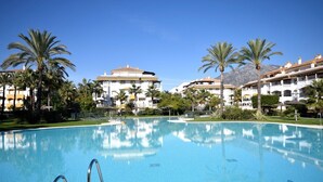 A swimming pool with clear blue water, surrounded by palm trees and white buildings with red-tiled roofs.