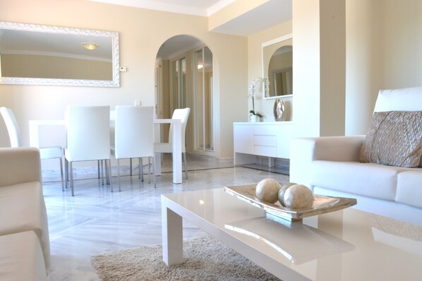 Un salon moderne avec un canapé blanc, une table basse agrémentée d’objets décoratifs, un coin repas avec des chaises blanches et un grand miroir au mur.