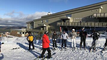 Un grupo de personas de pie en una ladera cubierta de nieve frente a un edificio con señalización de escuela de esquí y alquiler de equipos.