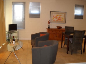 A living room with a TV on a glass table, two armchairs, a wooden cabinet, and a dining table with chairs.