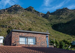 A small brick building with a tiled roof, large windows, and a chimney. It is situated in a hilly area with mountains in the background.