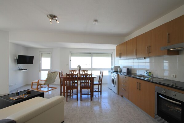A modern kitchen with wooden cabinets, a dining area with a table and chairs, a flat-screen TV mounted on the wall, and a built-in oven.