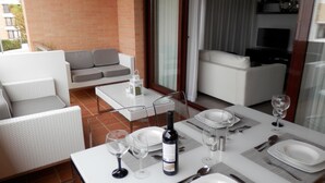 Un coin repas avec une table dressée pour deux personnes, comprenant des verres à vin, des assiettes et des couverts. On aperçoit un canapé et un téléviseur au fond.
