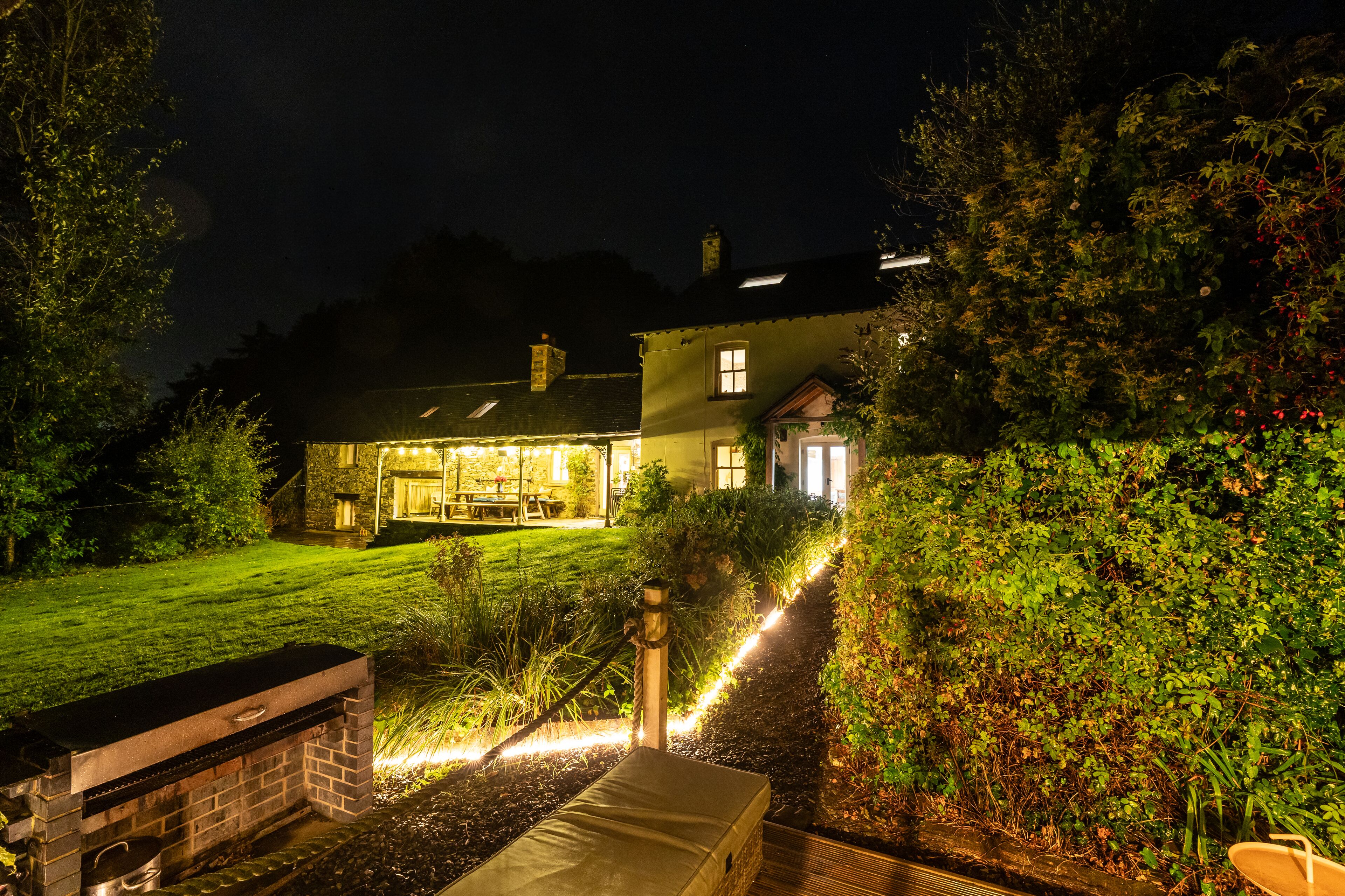 A house with a well-lit patio area, a barbecue grill, and a pathway leading to the entrance.