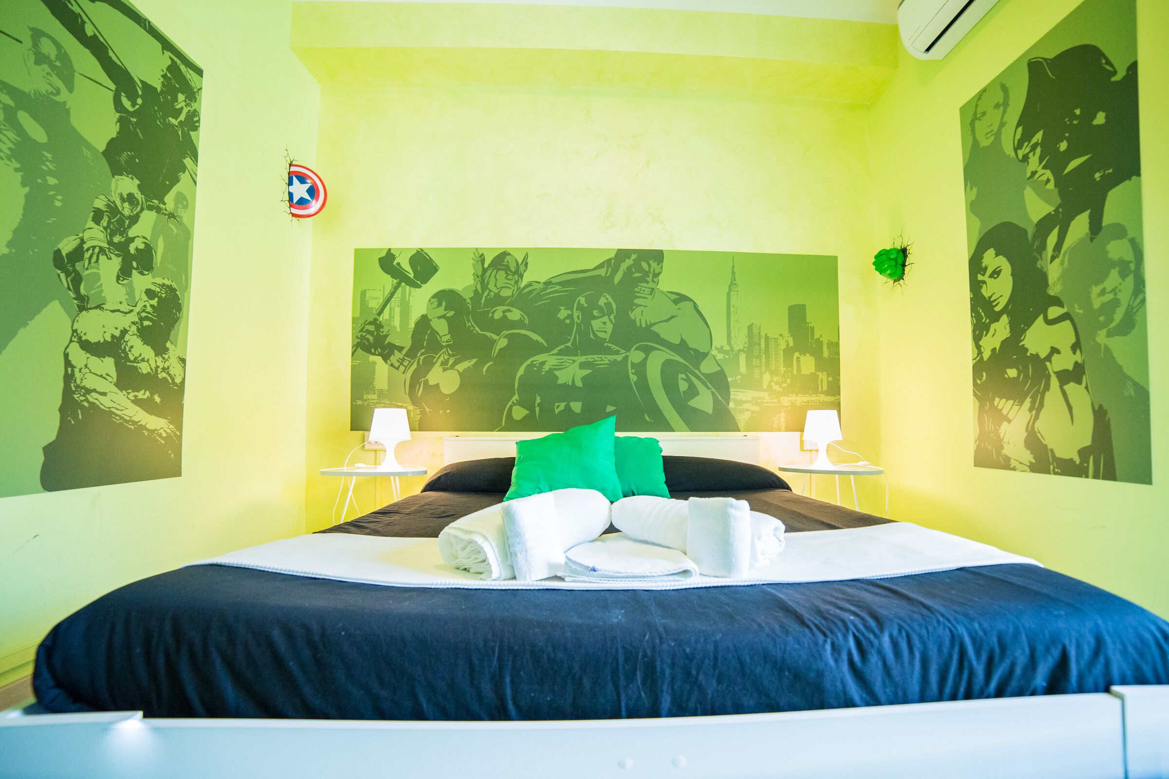 A hotel room with a bed, green walls, and superhero-themed artwork.