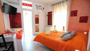 Ein Schlafzimmer mit orangefarbener Bettdecke, einem Schreibtisch mit Stuhl, einem Flachbildfernseher und einer Wand mit der Aufschrift 'Romantic Room'.