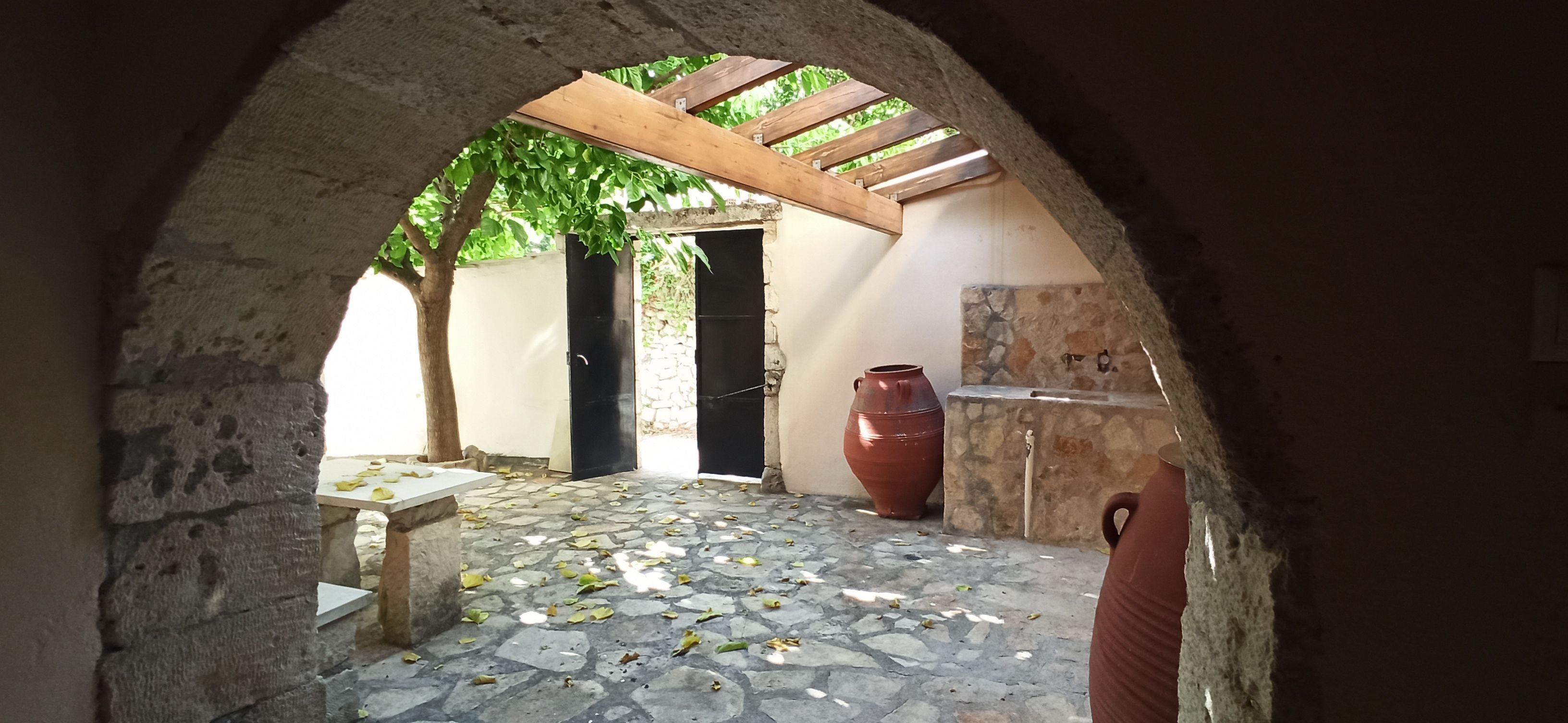 Un portail en pierre en arc de cercle mène à une cour intérieure dotée d’un sol carrelé, d’une pergola en bois, d’une porte noire et d’un pot rouge.
