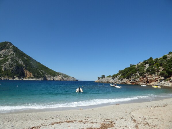 A beach with clear blue water, a sandy shore, and rocky hills in the background.