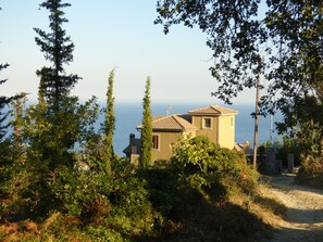 A coastal house with a tiled roof, surrounded by greenery and overlooking the sea.