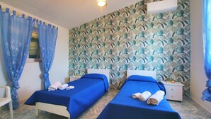 Une chambre avec deux lits, chacun recouvert d’une courtepointe bleue et doté d’une tête de lit blanche. On y trouve une fenêtre avec des rideaux bleus, un mur orné d’un motif végétal et un climatiseur fixé au mur.