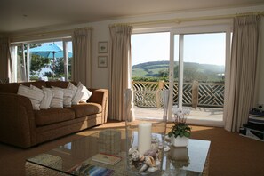 Sala de estar com sofá marrom, mesa de centro de vidro, um vaso com conchas e vista para uma varanda com guarda-corpo de madeira.