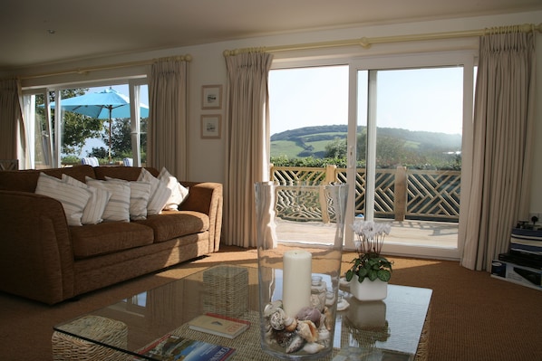 A living room with a brown sofa, a glass coffee table, a vase with seashells, and a view of a balcony with a wooden railing.