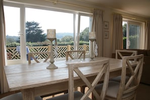 Uma mesa de jantar de madeira com cadeiras, vista para o exterior através de grandes janelas e uma varanda com guarda-corpo de madeira.