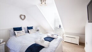 A bedroom with a sloped ceiling, a bed with blue and white bedding, a bedside table with a lamp, and a small white bench. - Flip Flops - Three Bedroom Cottage, Sleeps 6 (Crantock)