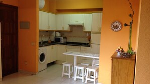 A compact kitchen with white cabinets, a washing machine, a microwave, and a dining area with a table and two stools.