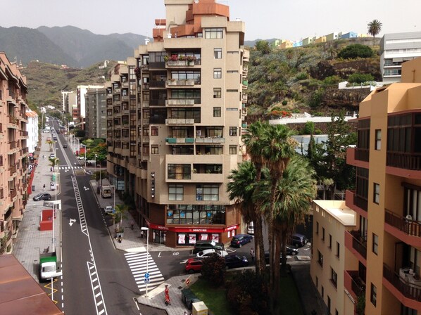 A multi-story building with balconies, a street in front, and a mountainous backdrop.