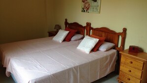 A neatly made bed with a pink patterned bedspread and several pillows, a wooden nightstand with a lamp, and a small wooden dresser.