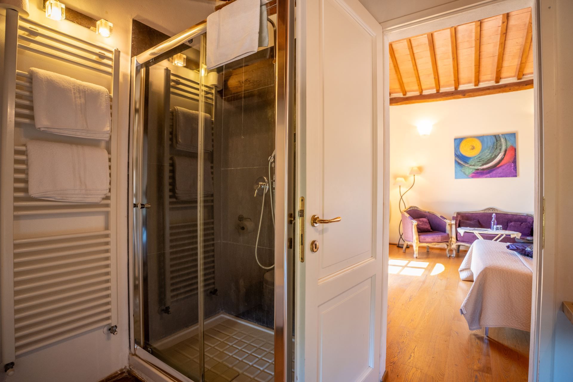 Camera d'albergo con cabina doccia in vetro, una porta bianca e vista su un soggiorno con divano e un quadro.