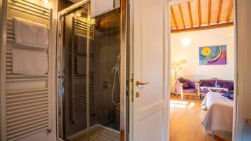 Camera d'albergo con cabina doccia in vetro, una porta bianca e vista su un soggiorno con divano e un quadro.