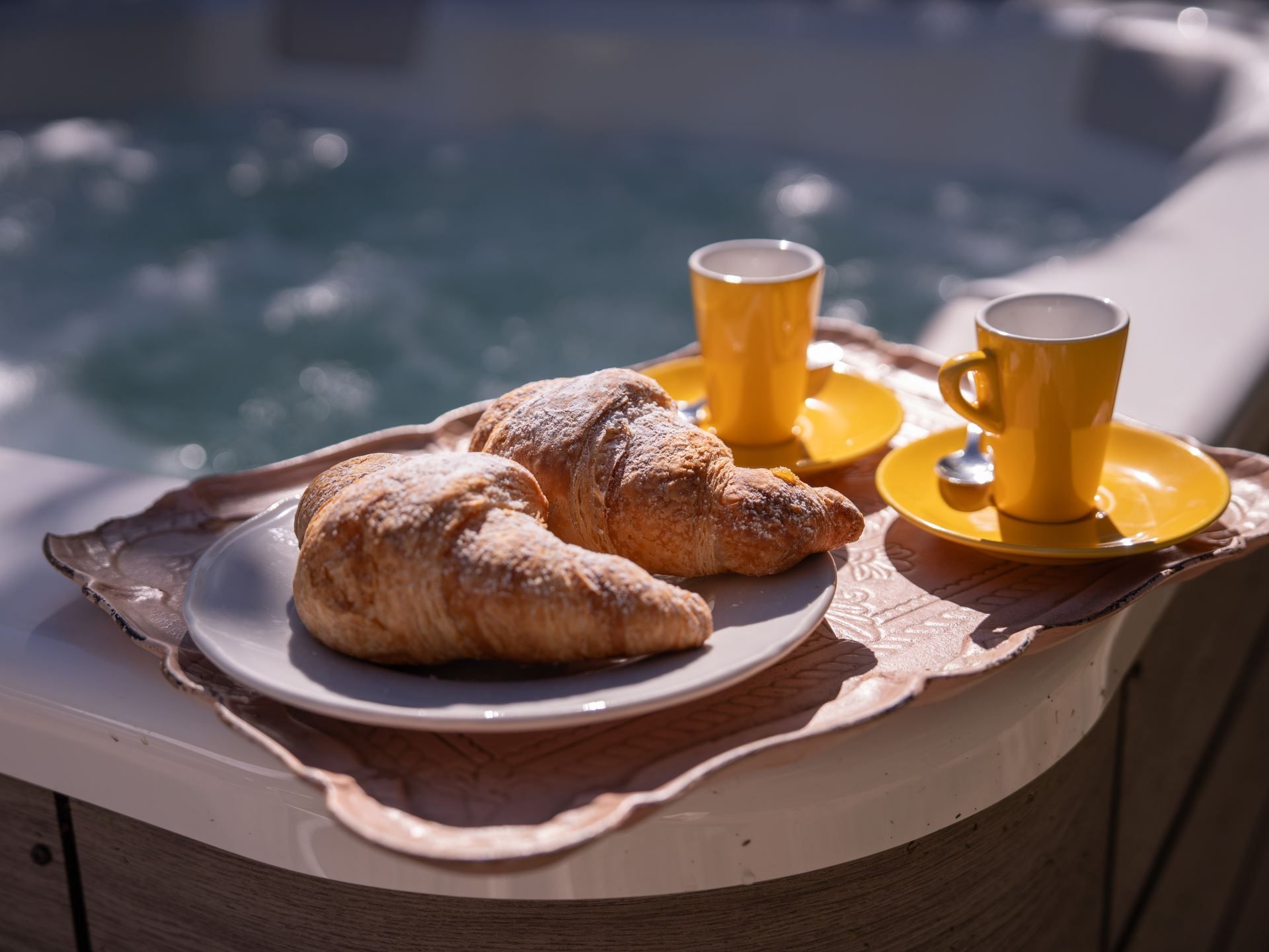 A tray with croissants and yellow cups on a wooden table near a pool.