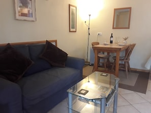 Una sala de estar con un sofá azul, una mesa de centro de vidrio, una mesa de comedor con sillas y una lámpara de pie.