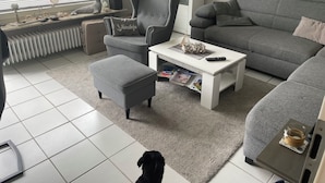 Una sala de estar moderna con un sofá y un sillón grises, y un banquillo a juego. En el centro, una mesa de centro blanca con revistas y un control remoto. Un radiador está debajo de la ventana, y se ve una pequeña estantería con objetos decorativos.