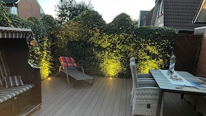 Una terraza de madera con una silla de descanso, una mesita con una lámpara y un seto al fondo.