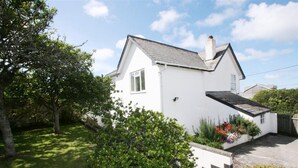 A white two-story house with a sloped roof, surrounded by lush greenery and a small garden with colorful flowers. - Clovelly Cottage - Four Bedroom Cottage, Sleeps 8 (Crantock)