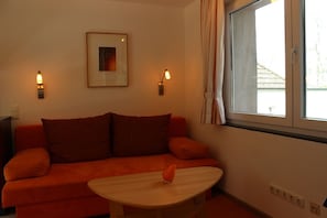 A room with a red sofa, a small wooden table, two wall-mounted lamps, a framed picture, and a window with curtains.