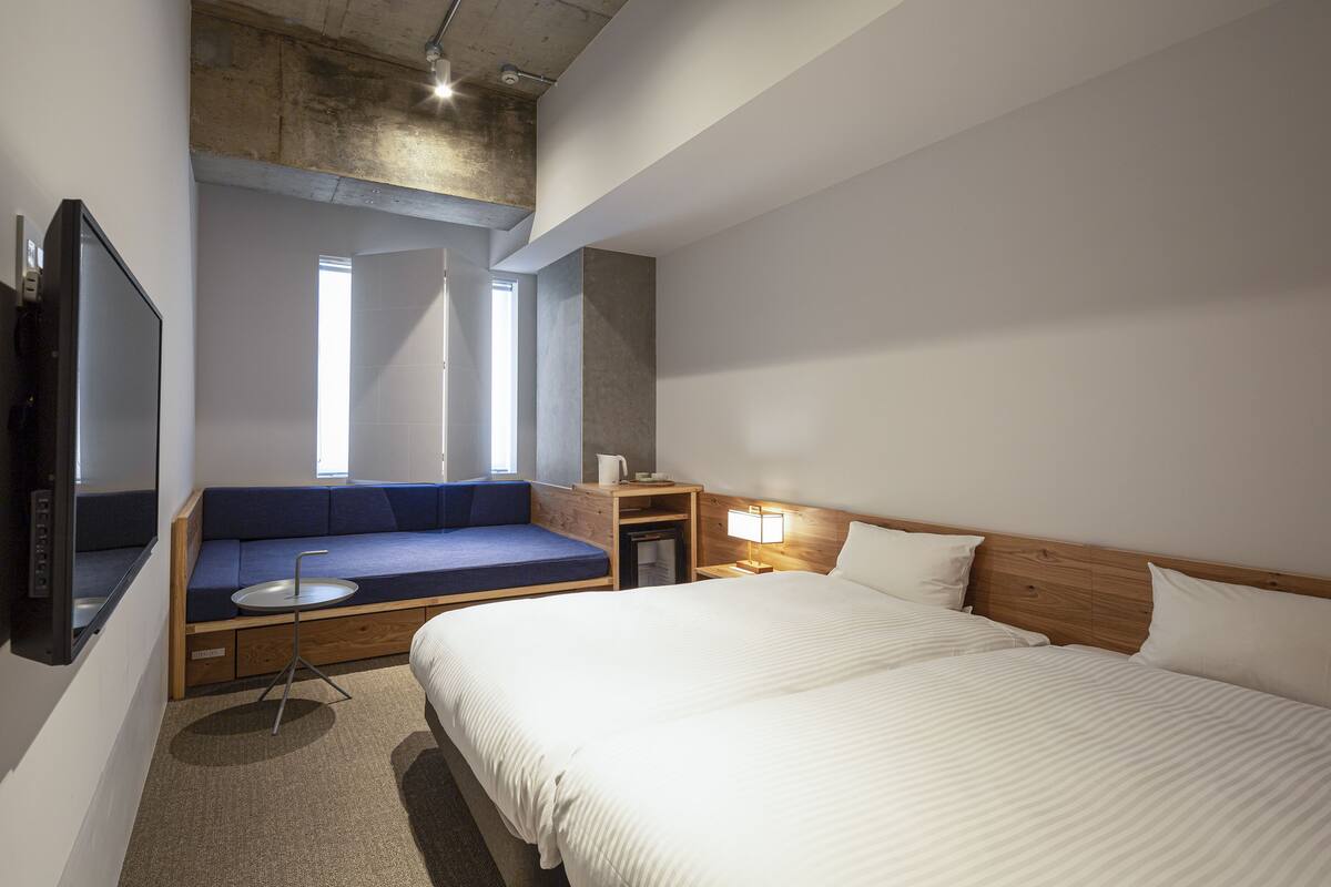 Ein modernes Hotelzimmer mit einem großen Bett, einer Schlafcouch, einem Fernseher, einem Nachttisch mit Lampe und einem Fenster mit Tageslicht.