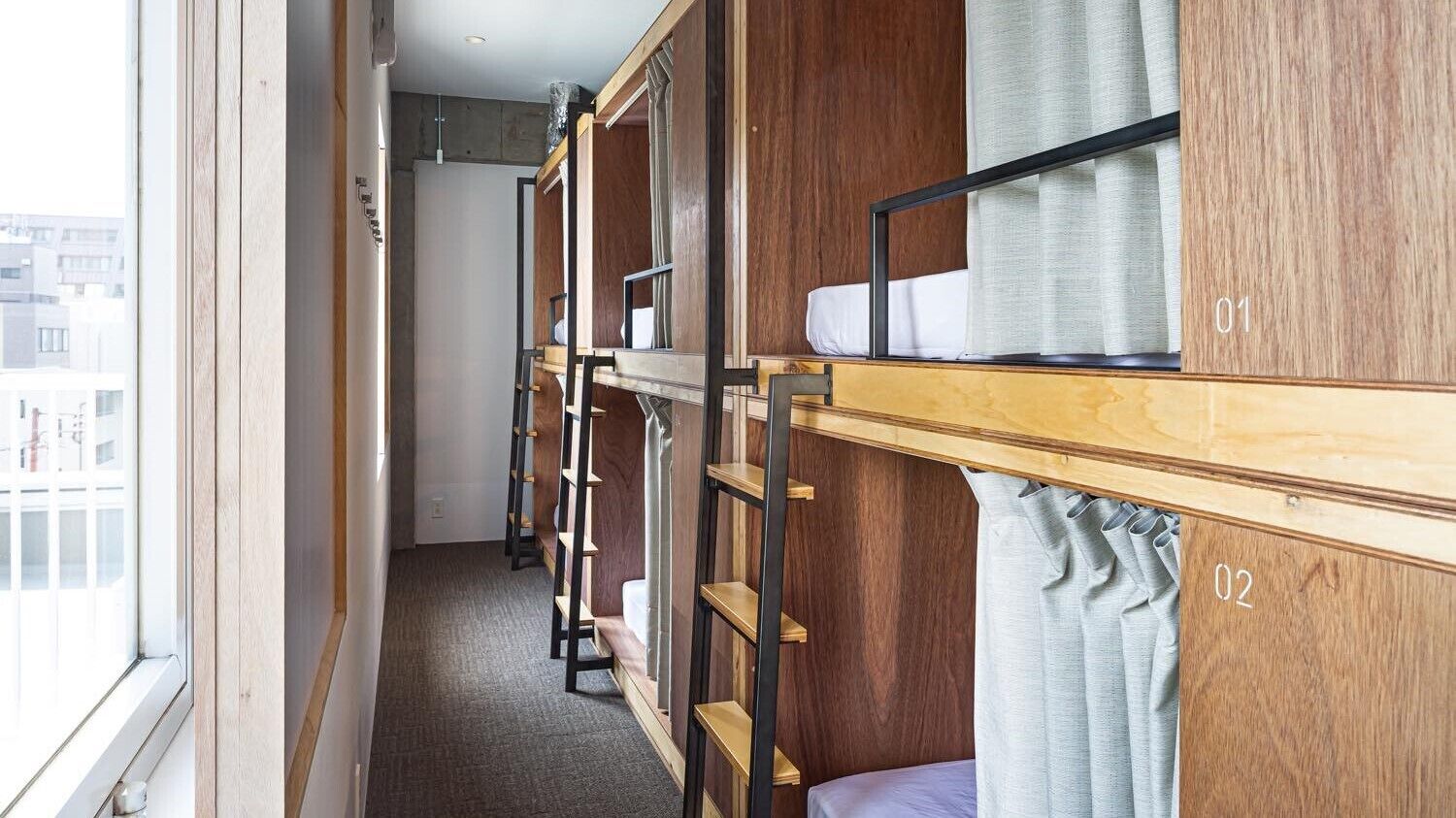 A narrow corridor with bunk beds, numbered 01 and 02, wooden walls, and a window with a city view.