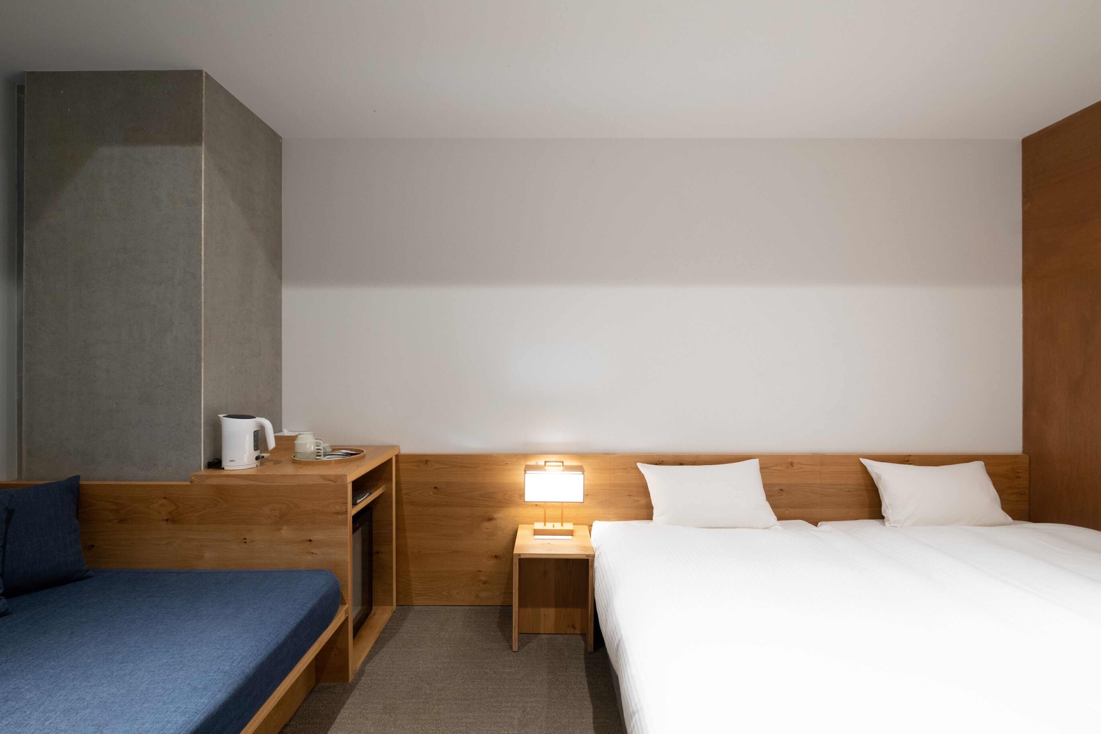 A hotel room with a bed, a small bedside table with a lamp, a coffee maker, and a blue sofa.