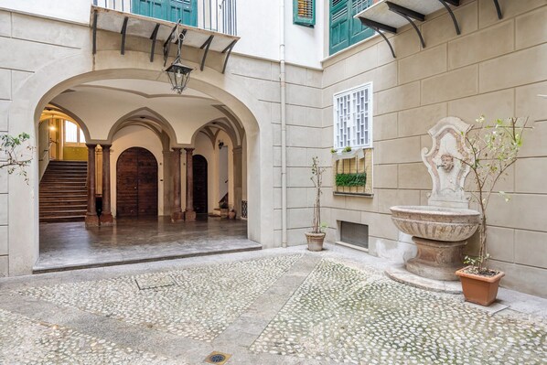 An indoor courtyard with stone flooring, a central fountain, and a staircase leading to an upper level.