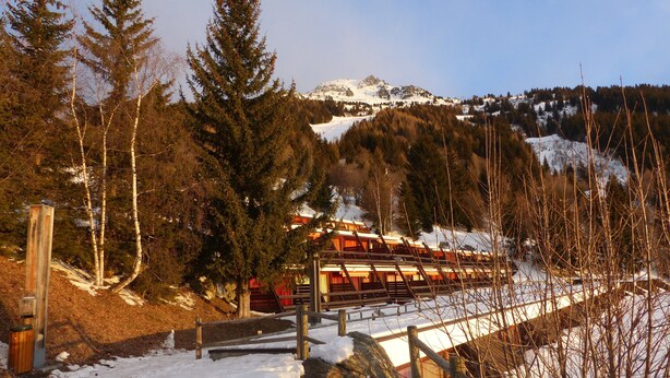 Un espacio al aire libre cubierto de nieve, con bancos de madera, una valla de madera y un edificio con tejados rojos al fondo.
