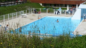 Una piscina rectangular con un deck de madera alrededor, un área de estar cubierta y un edificio al fondo.