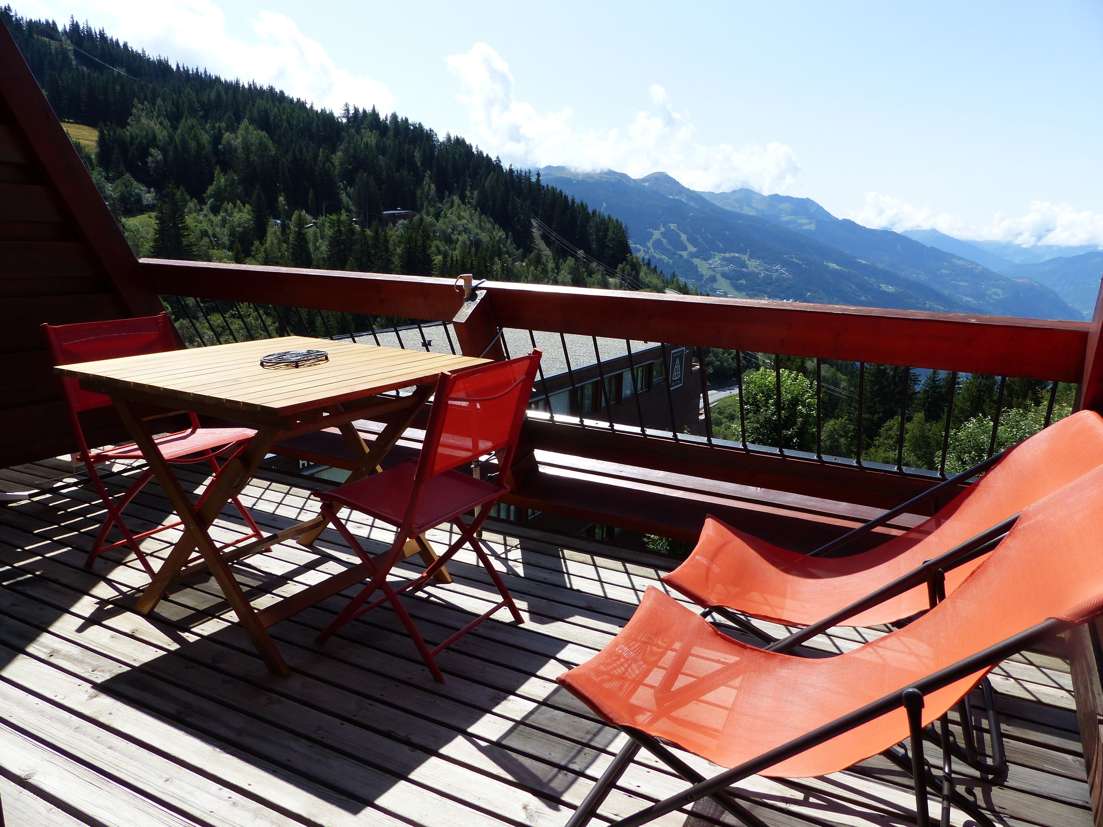 Una terraza de madera con una mesa y sillas rojas, con vistas a un paisaje montañoso.
