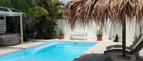 A swimming pool with a thatched roof, surrounded by palm trees and potted plants.
