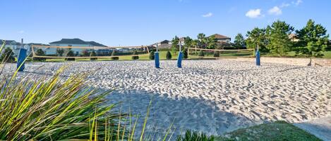 A sandy beach volleyball court with blue volleyball nets and posts, surrounded by greenery and residential buildings in the background.
