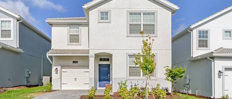 A two-story residential building with a blue front door, white garage doors, and a small tree in the front yard.