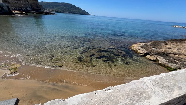A coastal scene with clear water, rocky shoreline, and a building with multiple windows.
