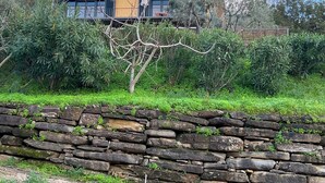 A stone wall in front of a yellow building with a chimney, surrounded by greenery.