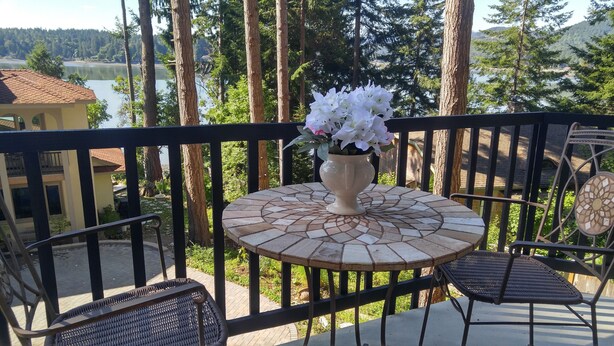 A balcony with a round table and chairs, a vase with flowers, and a view of a lake and forest.