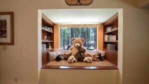 A cozy reading nook with a large teddy bear centerpiece, surrounded by bookshelves and a window with a forest view.