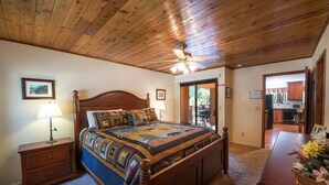 A bedroom with a wooden ceiling, a large bed with a colorful quilt, a nightstand with a lamp, and a view into the kitchen through an open door.