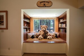 Un rincón acogedor para leer con un gran osito de peluche como centro, rodeado de estanterías y una ventana con vistas a un bosque.